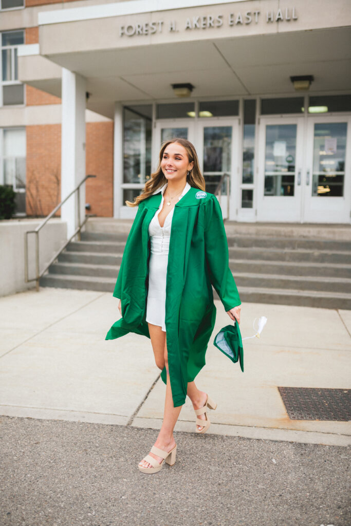 A brunette, graduate wearing a white dress under her green graduation gown walking outside of Forest Akers East Hall on Michigan State University Campus