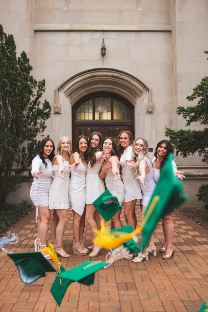 A group of friends wearing white dresses, throwing their caps toward the camera and smiling at Beaumont Tower on Michigan State University campus.