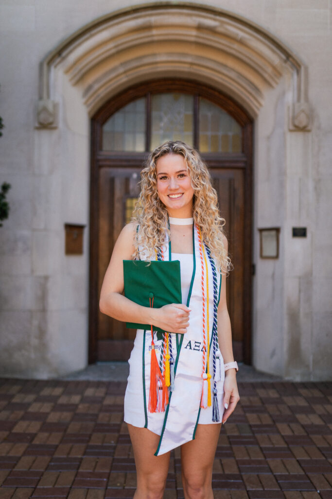 A blonde graduate wearing a white dress, her graduation tassels and holding her green graduation cap at Beaumont Tower on Michigan State University campus