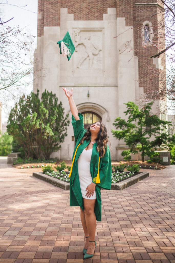 A brunette graduate wearing her tassels and green graduation gown, throwing her cap in the air at Beaumont Tower on Michigan State University Campus