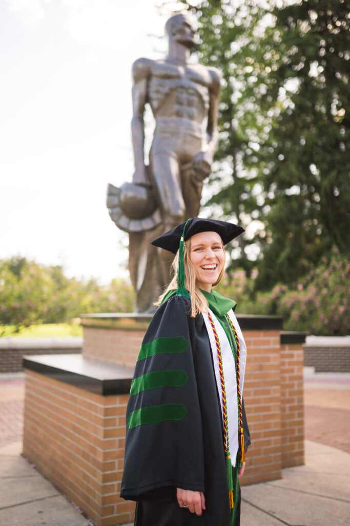 A blonde doctoral graduate on a sunny day at the Sparty Statue. She is wearing full doctoral graduation cap, gown and tassels. 