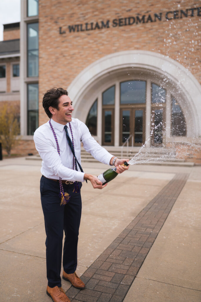 A brunette graduate wearing a white shirt his graduation tassels and black pants, spraying a bottle of champagne at the William Seidman Center on Grand Valley State University campus in Grand Rapids