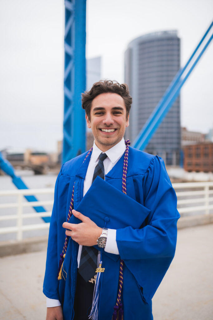 A brunette graduate wearing his blue Grand Valley State University gown and tassels and holding his graduation cap on a bridge in Grand Rapids