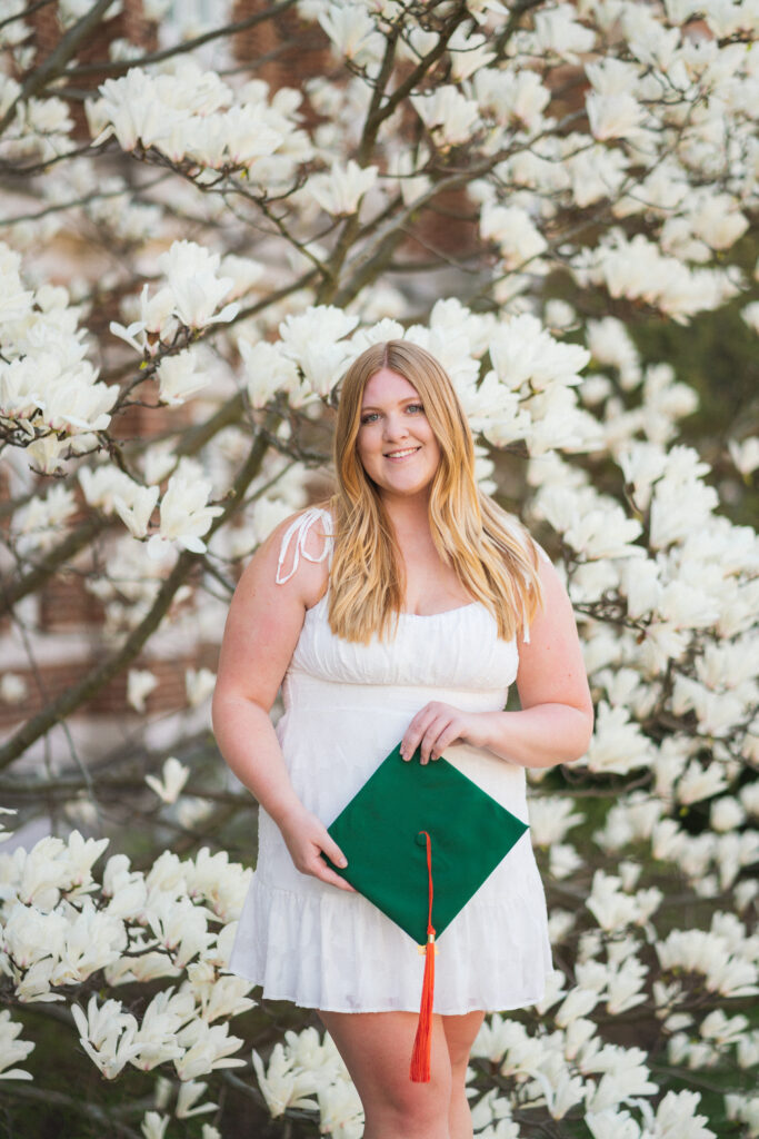 A blonde graduate holding her graduation cap, wearing a white dress in front of a flowering white tree on Michigan State University campus.